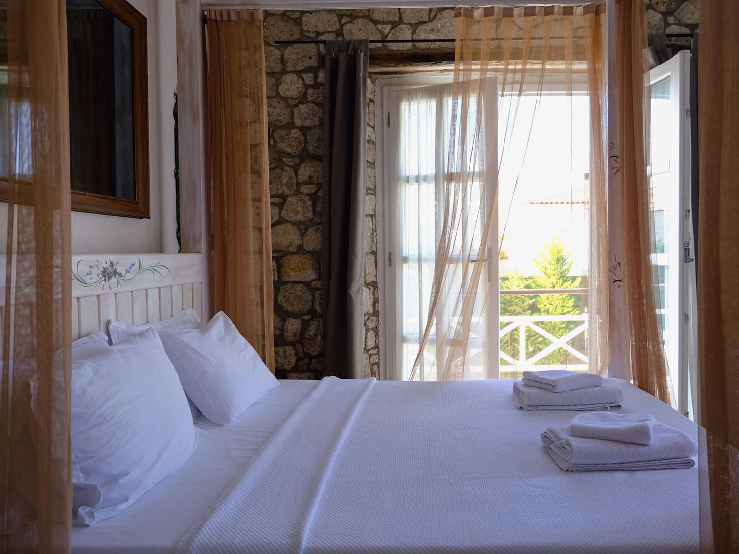 Cozy tropical bedroom with white linens and soft curtains, sunlight streaming through open balcony doors with garden views.