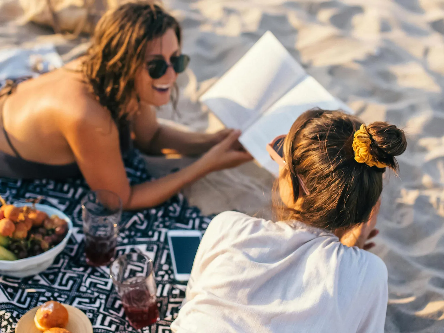 Two women enjoying a relaxed picnic on the sandy beach with fruit, drinks, and a book under the Puerto Rico sun.