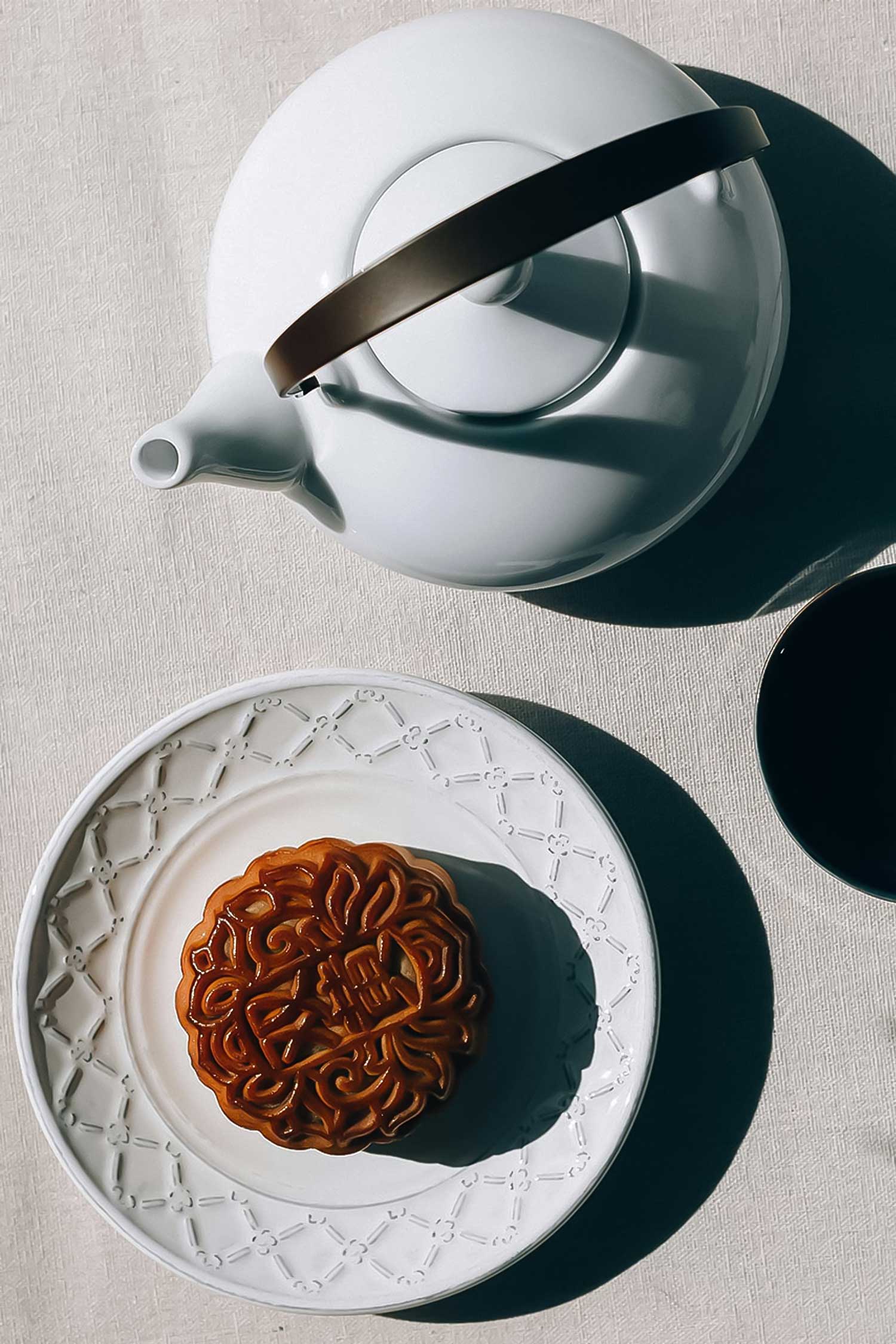 Overhead view of a breakfast table with teapot, cup, and pastry, sunlight casting soft shadows on the linen.