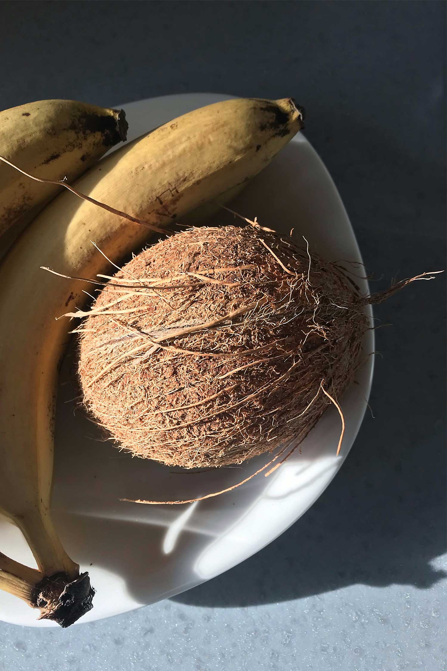 Fresh coconut and bananas on a white plate, captured in warm natural light on a stone surface.