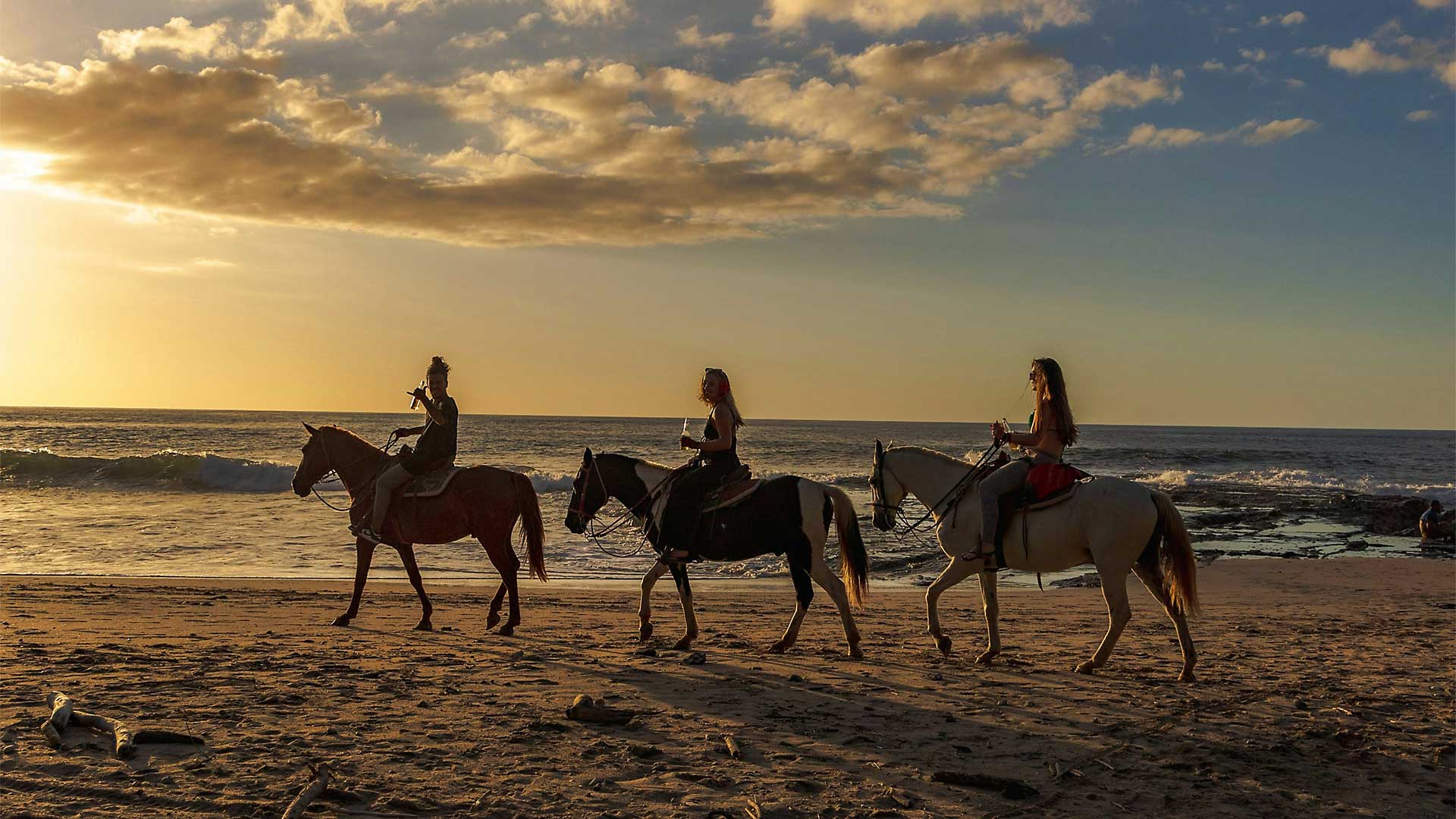 Three people horseback riding along the sandy Puerto Rico beach at sunset with waves in the background.