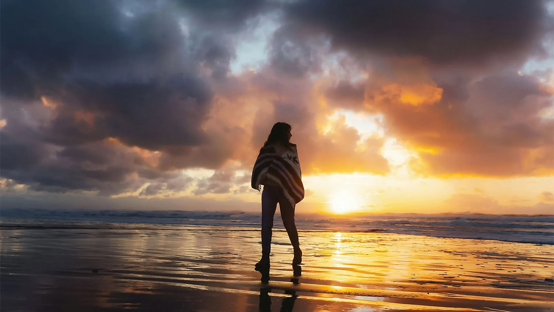 Woman walking on the beach wrapped in a blanket at sunset, with dramatic clouds reflecting on wet sand.
