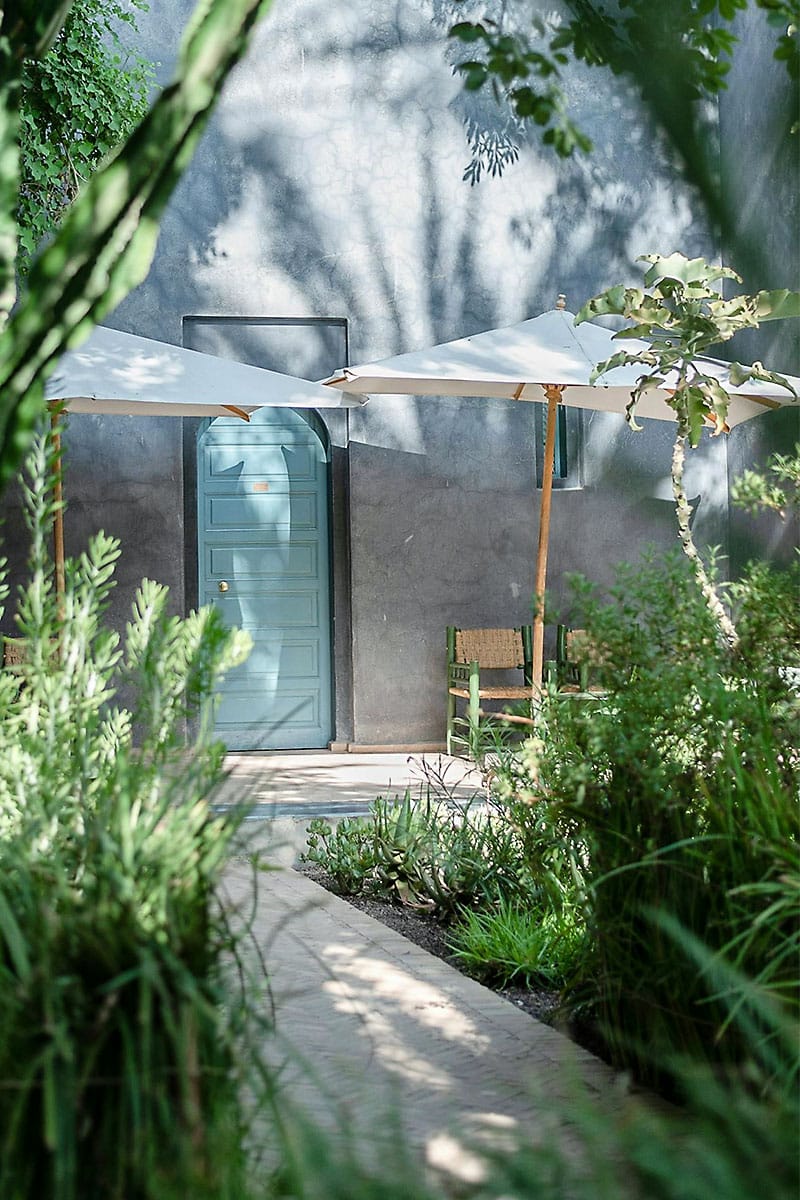 Tropical greenery framing a hacienda-style blue door with outdoor seating and umbrellas at Coquí Hideaway in Puerto Rico.